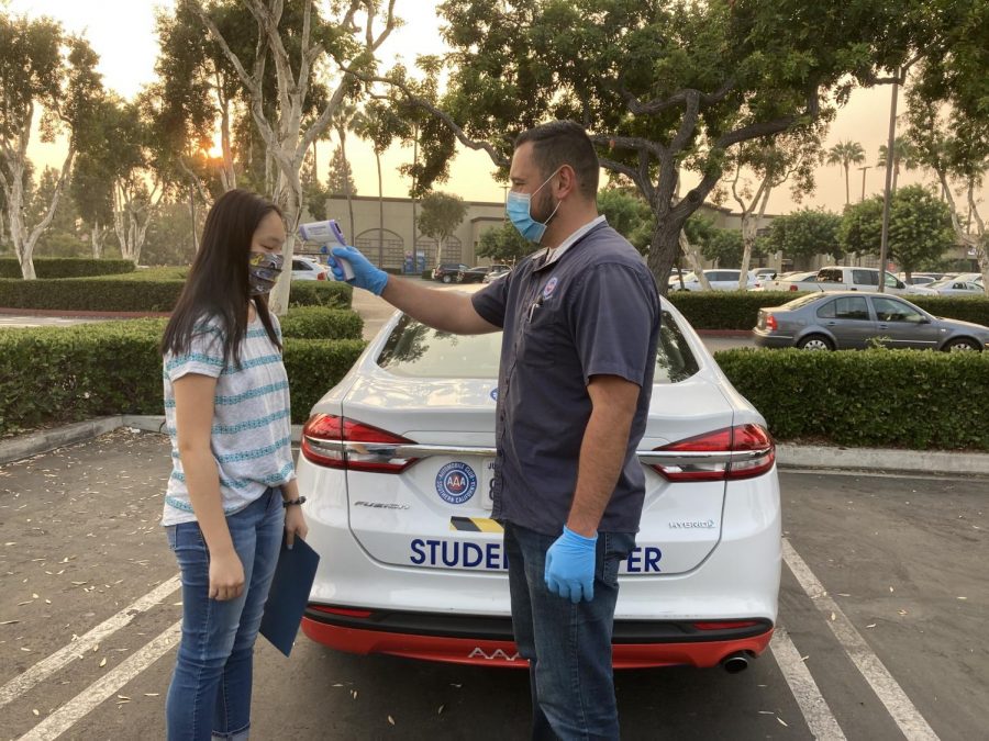 Before her Sept. 15 AAA driving lesson, senior Hope Li (left) dons a face mask as her instructor, Daniel Pedraza, takes her temperature in front of the AAA student driver car. Li has been taking lessons since July, following coronavirus precautions like wearing gloves in the car and sitting on a plastic seat cover.