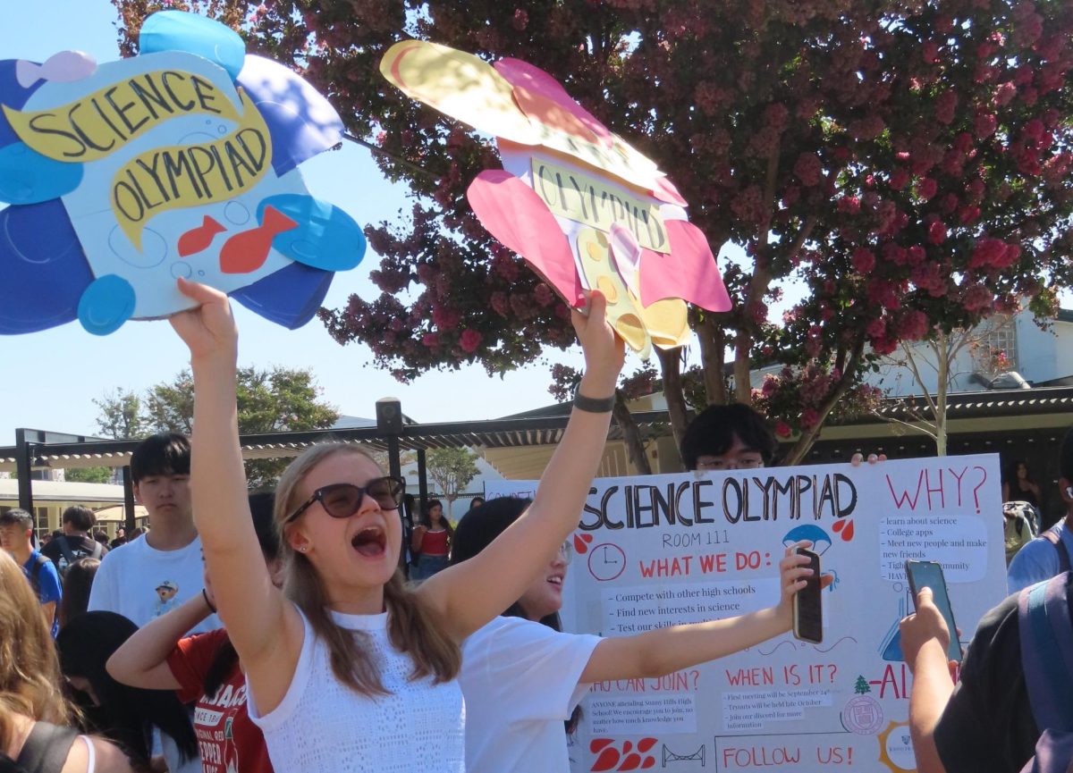 Holding up two Science Olympiad signs, senior Sophia Kiker tells students to join her group. Kiker, one of the club's presidents, was among the two other Science Olympiad captains at their table to promote what they do during break in the quad on the second day of Club Rush on Wednesday, Sept. 4.