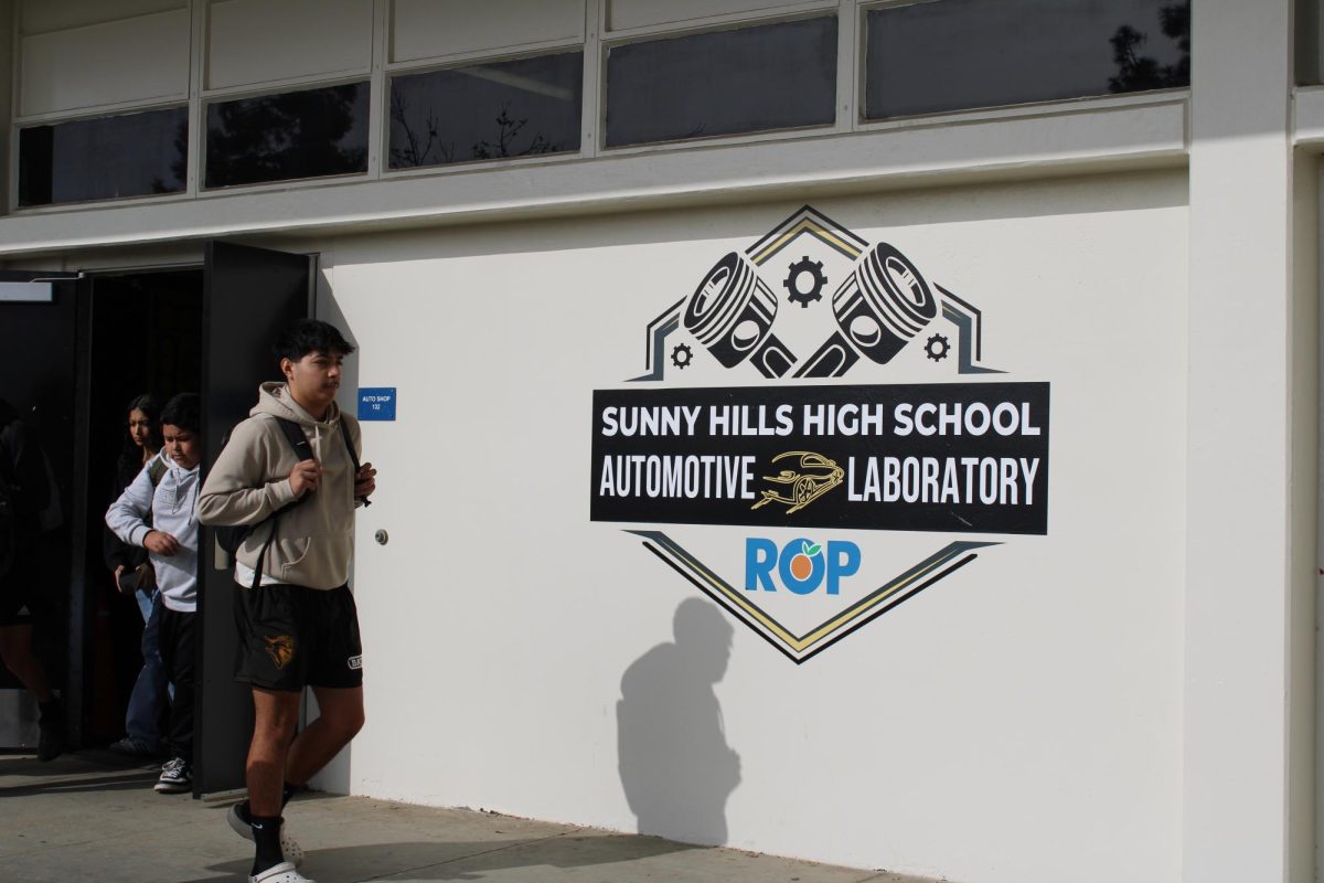 Students pass by the new auto tech program sign – created by then-senior Cali Watson – that features two car pistons in the shape of an “X” above a black rectangular sign with the text “Sunny Hills High School” on top of “Automotive Laboratory.” A car outlined in gold splits the words “Automotive” and “Laboratory” while the ROP [Regional Occupational Program] logo rounds out the design at the bottom.