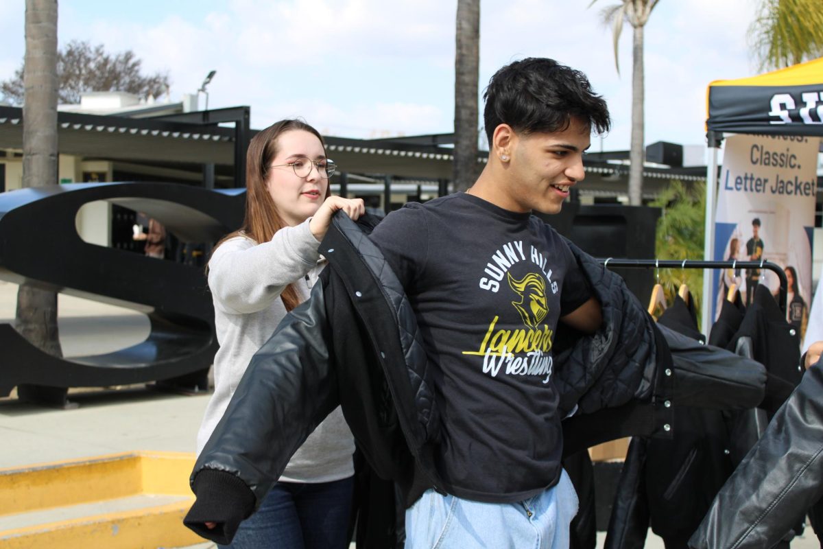 Sophomore Aarav Arora (right) tries on a Jostens senior letter jacket with the help of an employee during lunch on Tuesday, Feb. 11, in the quad. The Minnesota-based company sent its representative to showcase Jostens’ high school memorabilia, which included class rings and other attire for sale. Those visiting the display tent were eligible to spin a wheel to win random prizes ranging from candy to class tassels.