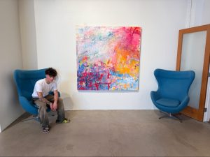 Senior Jasper Hanrahan sits next to his displayed painting at his Downtown Fullerton art gallery on Friday, Aug. 29, a day before the grand opening.